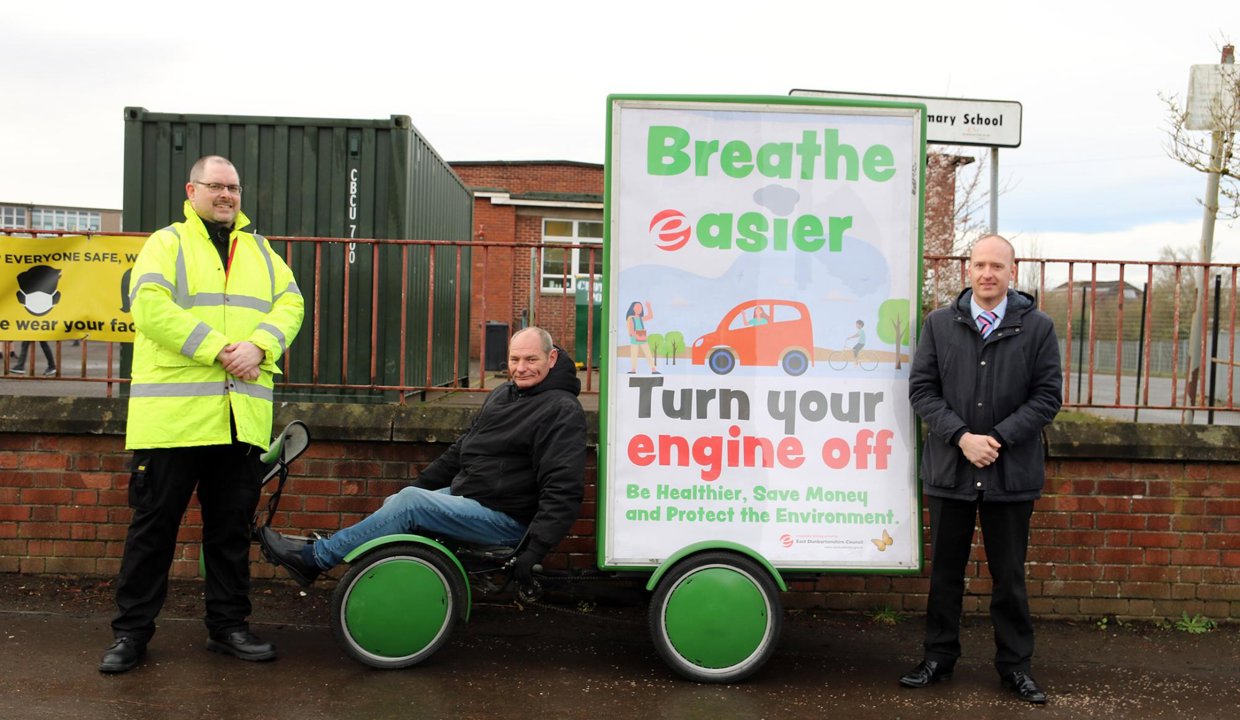 Three men pictured outside a primary school beside the ad-bike trailer showing the 'breathe easier turn your engine off' poster.