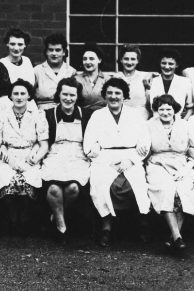 A black and white image depicting two rows of woman dressed in white work overalls and seated and posing for a photograph