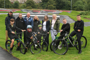 A group of people some standing and others on the bikes. Behind them is the pump track.