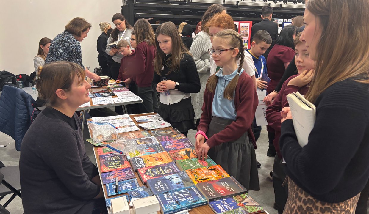 Two busy book stalls with tables covered in books, with people standing looking at the merchandise and talking with the women sitting behind the tables. 