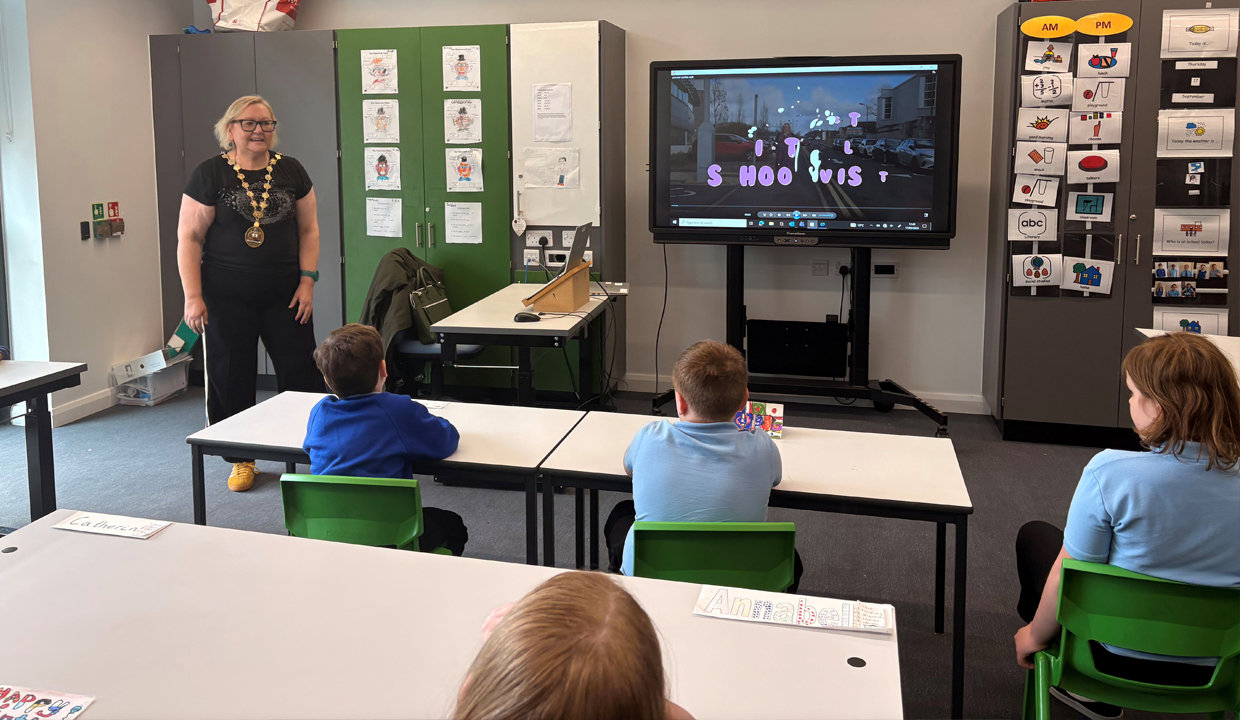 Provost Renwick standing in a classroom at Woodland View School in front of a class of pupils sitting at their desks.  To her left is a tv screen where she is showing her video to the pupils. 