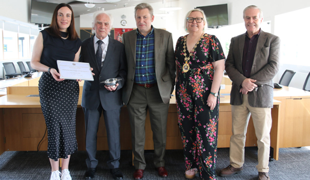 Five smartly dressed people standing in a row.  Woman to the left is holding a framed certificate and the man next to her is holding a silver quaich. 