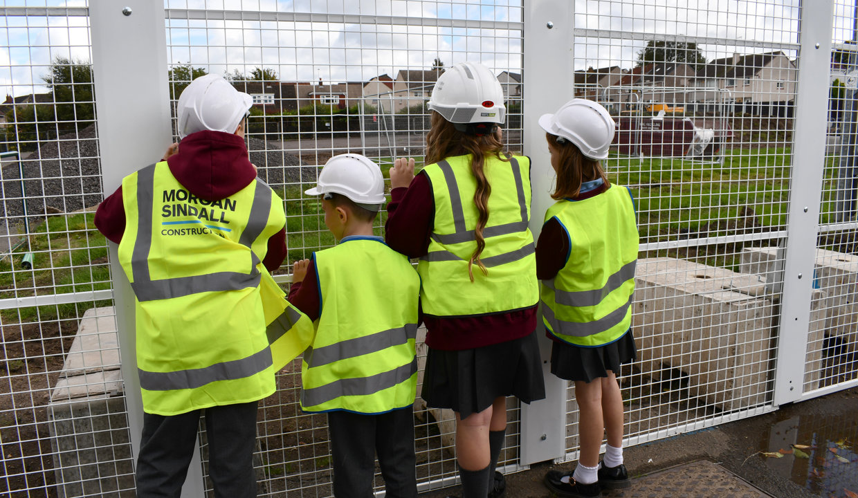 Four children wearing hi vis vests and hard hats looking through gapped fencing onto a building site.