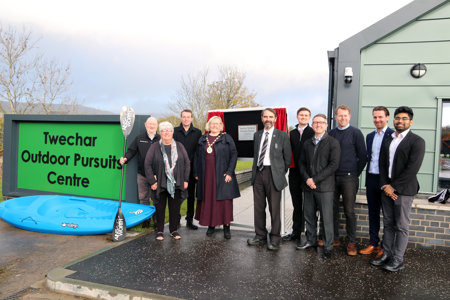 A group of officials, including East Dunbartonshire Provost and Council Leader, stand in a front of a one storey building next to a large sign which reads Twechar Outdoor Centre. Some canoes are propped up next to the sign. 