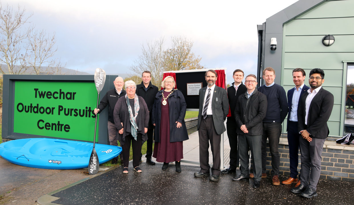 A group of officials, including East Dunbartonshire Provost and Council Leader, stand in a front of a one storey building next to a large sign which reads Twechar Outdoor Centre. Some canoes are propped up next to the sign. 
