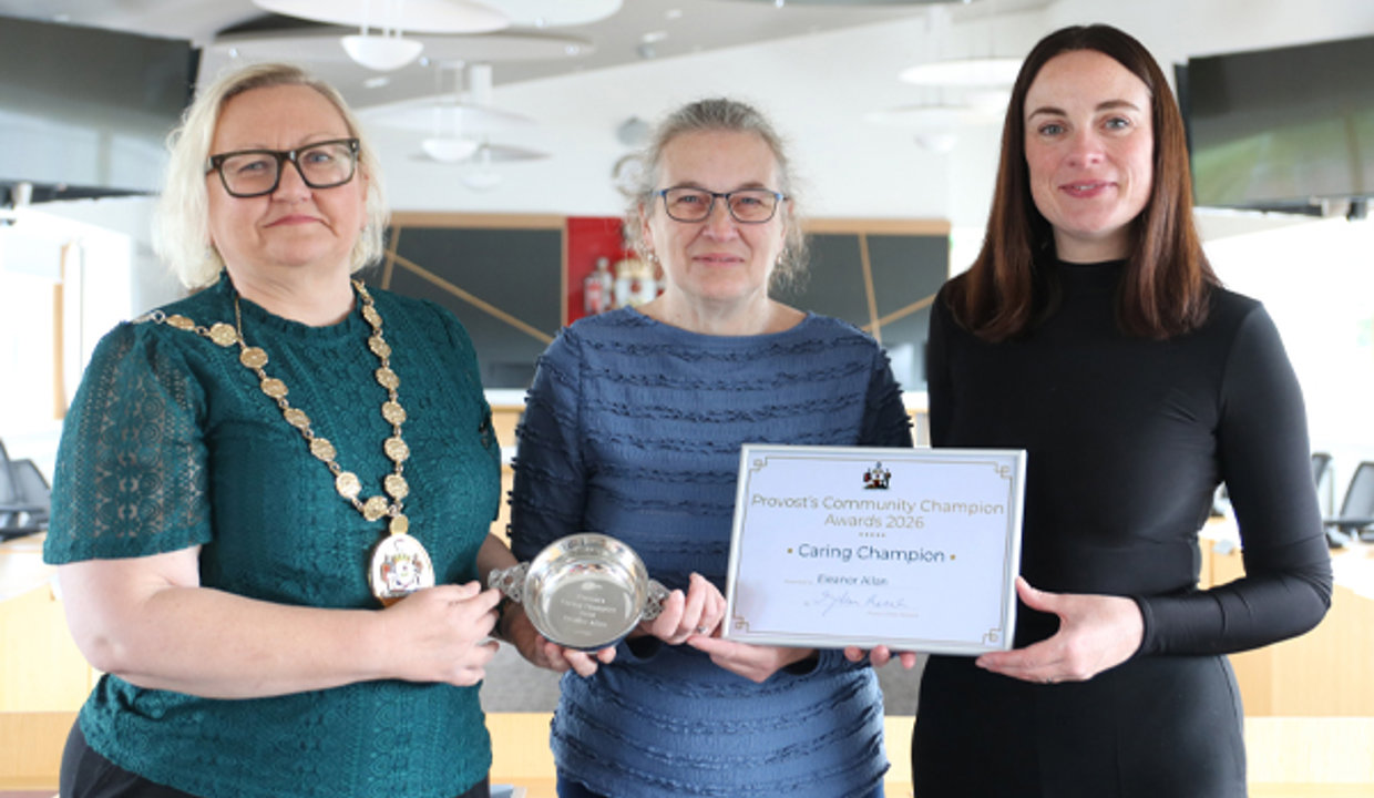 Three women standing together in Council chambers.  The woman on the left is wearing a chain of office and holding a silver quaich.  The other two women are holding a framed certificate between them. 