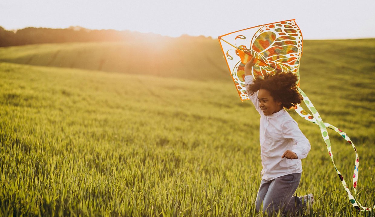 Child running through a grassy field holding a kite with the sun setting in the background