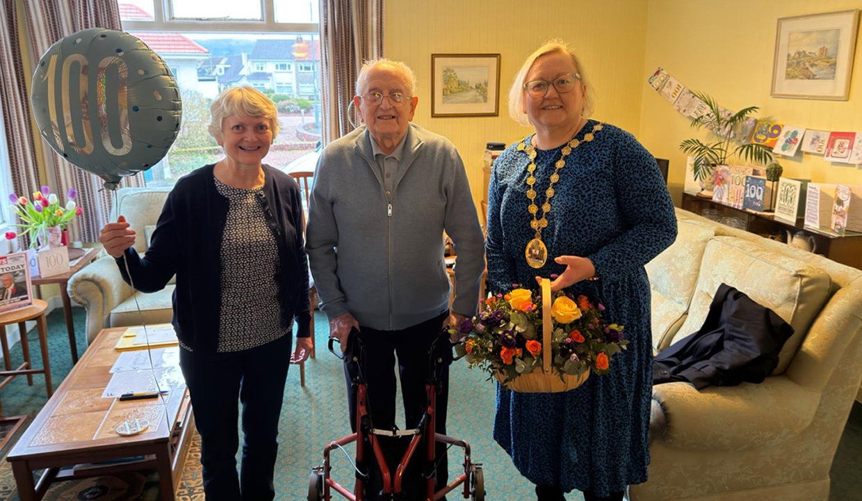 Image of three people standing in bright lemon painted living room, with matching lemon suite. The lady on the far left is holding a blue ‘100’ balloon in her hand.  An older gentleman is standing in the middle with a walking aid and to his right is woman in a blue dress, wearing chains of office and holding a basket of beautiful flowers. 