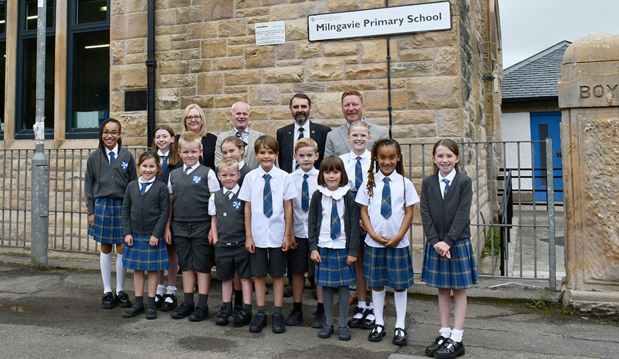 Group of adults and school pupils standing outside blond sandstone school building with a sign which reads Milngavie Primary School. 