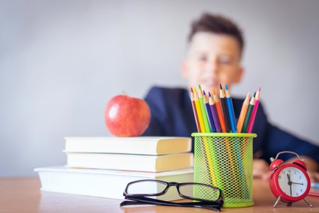 Pile of books, pair of glasses, tub of pencils and alarm clock on a table with a boy sitting in the background