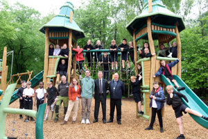 A group of around 20 school children pose on a climbing frame which has a rope bridge and two wooden turrets at either end. A group of five adults stands in the middle of the children. 