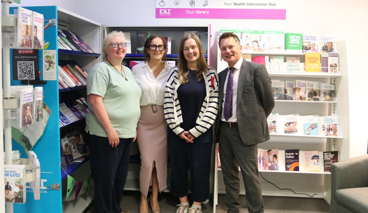 A man and three women stand in front of shelving which displays a variety of leaflets and books on health topics. Above them is a sign which reads Health Information Hub. 
