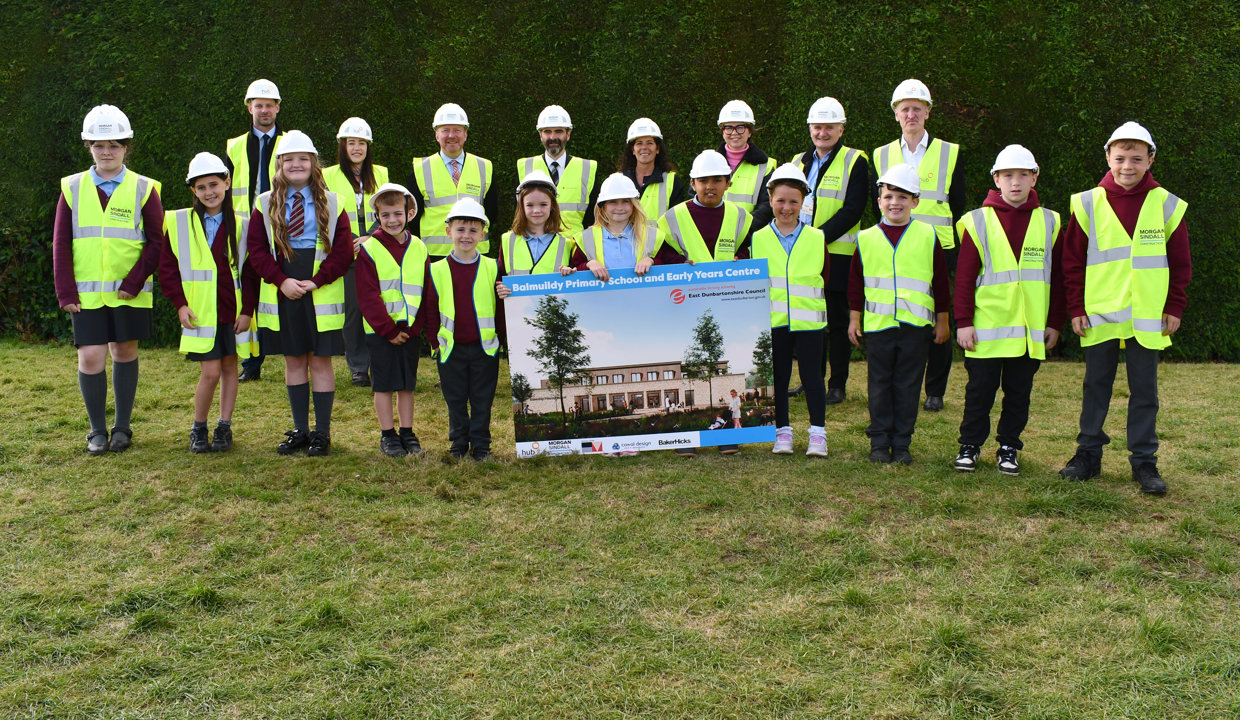 Adults and children wearing hi vis vests and hard hats standing on grass.  A green hedge is behind them. In the foreground children are holding a large image of a new school. 