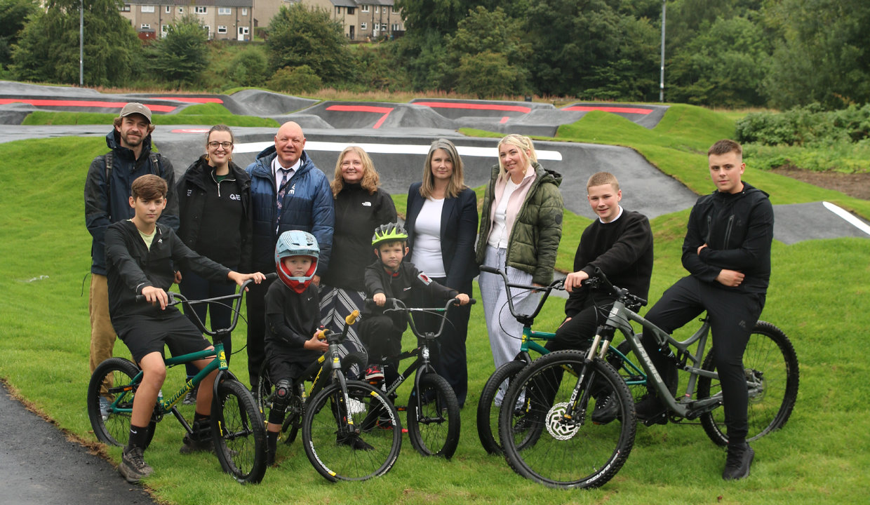 A group of people some standing and others on the bikes. Behind them is the pump track.