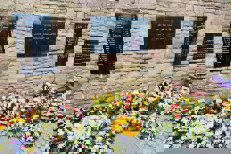 Beautiful, colourful flower arrangements laid against a sandstone-coloured wall with large black plaques interspersed along the brick work.  The plaques contain a list of names of those who died at the Auchengeich Mining Disaster in 1959.  