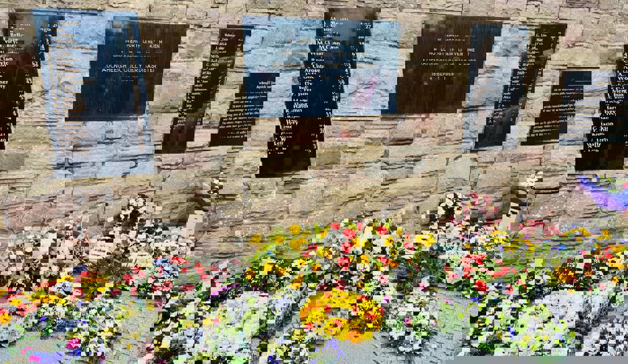 Beautiful, colourful flower arrangements laid against a sandstone-coloured wall with large black plaques interspersed along the brick work. The plaques contain a list of names of those who died at the Auchengeich Mining Disaster in 1959.