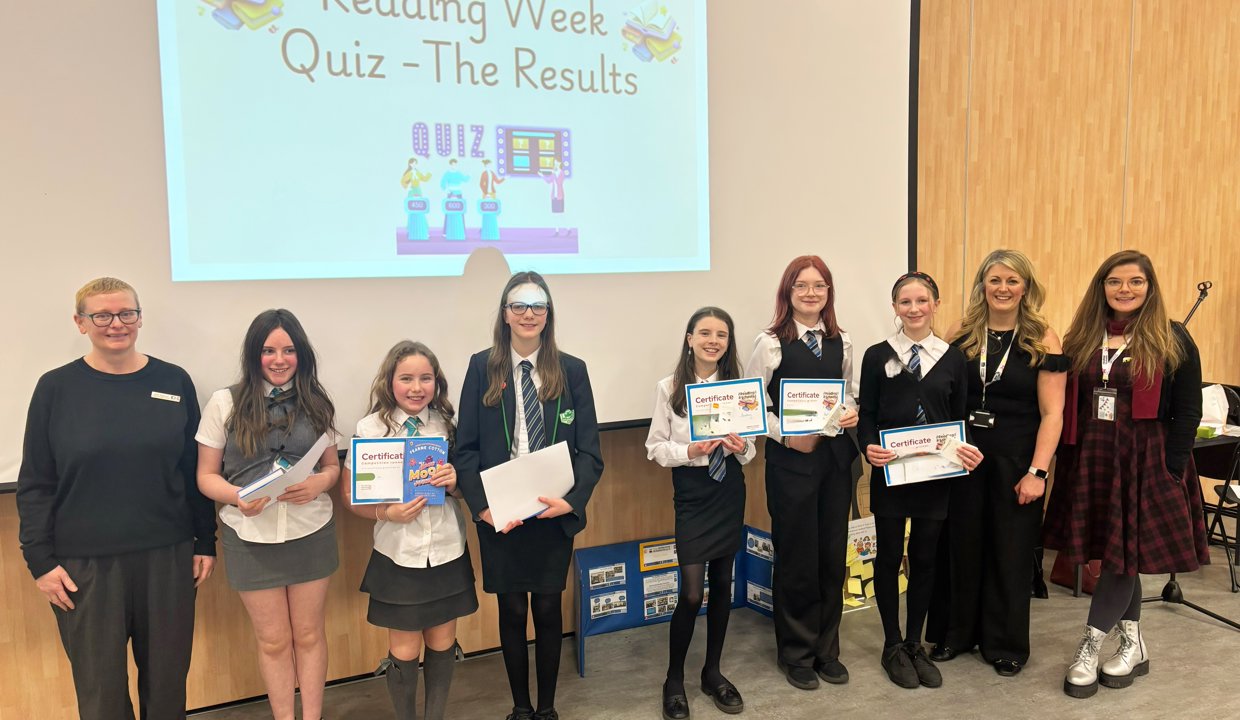 Group of adults and children wearing school uniform and holding certificates standing in front of a white projector screen. Projected onto the screen are the words ‘Reading Week – Quiz – The Results’ 