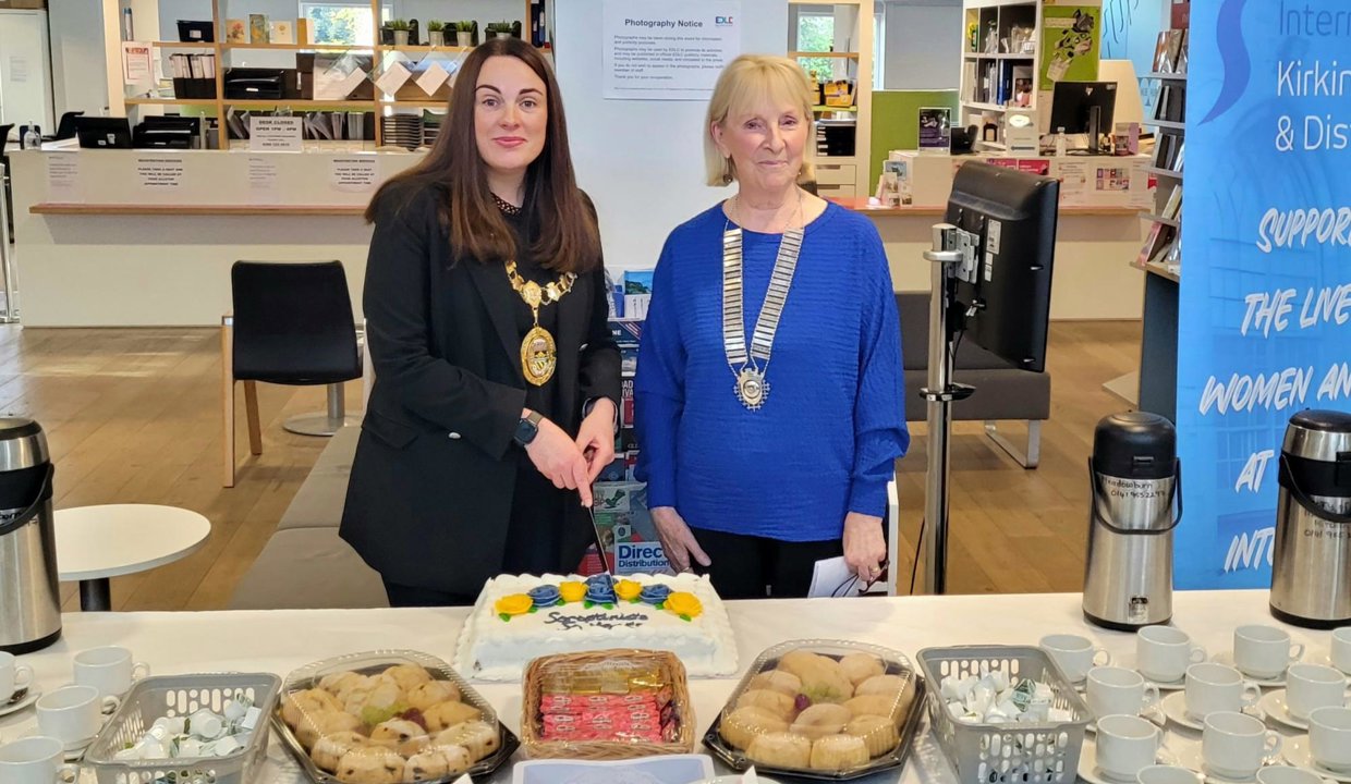 Depute Provost Colette McDiarmid and Soroptimist standing behind a table covered in baked goods and cutting a decorated cake.