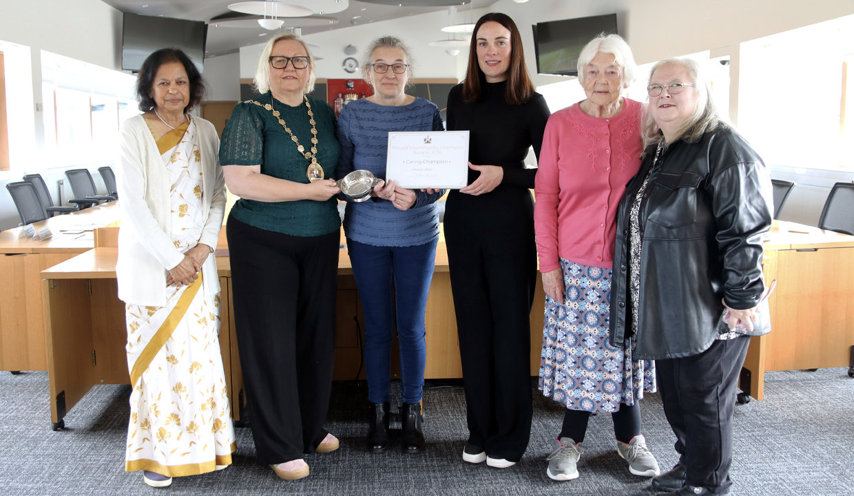 Formal shot of six women standing in Council chambers.  The women in the centre of the pic are holding a framed certificate and a quaich. 