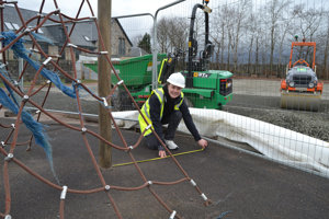 Girl wearing hi vis vest and hard hat measuring the ground in a playground. In the foreground is climbing apparatus and behind her is a green tractor and red roller vehicle. 
