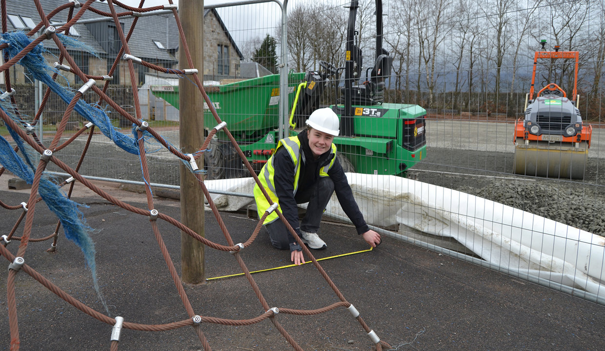 Girl wearing hi vis vest and hard hat measuring the ground in a playground. In the foreground is climbing apparatus and behind her is a green tractor and red roller vehicle. 