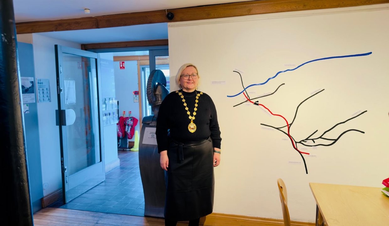 Woman dressed in black top and black leather skirt and chains of office, standing in front of a train route map.  A table and chair is in the front right of the picture and in the rear left is an open door. 