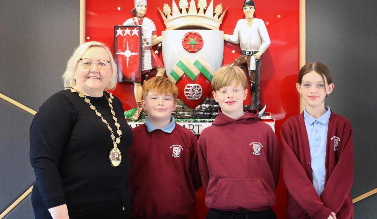 Provost Renwick, Ruairidh, Isaac and Sophia standing in front of East Dunbartonshire’s Coat of Arms which is mounted on the wall. Crest has red background, with a shield in centre and soldier and a miner at either side of coat of arms.
