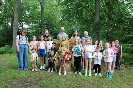 a group of people stand round a wooden statue of a fairy