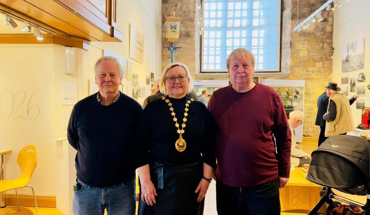 Three people standing in a large building that looks like a church.  Behind them is a large arched window and a brick work wall.  The men stand on either side of a woman wearing black clothing and chains of office. 