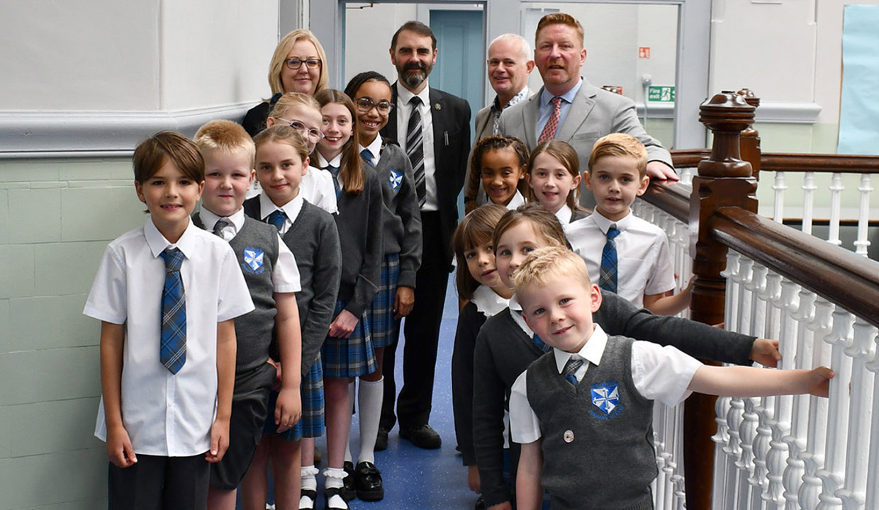Group of adults and primary school aged children standing against a banister in a school.  Children are wearing school uniform.