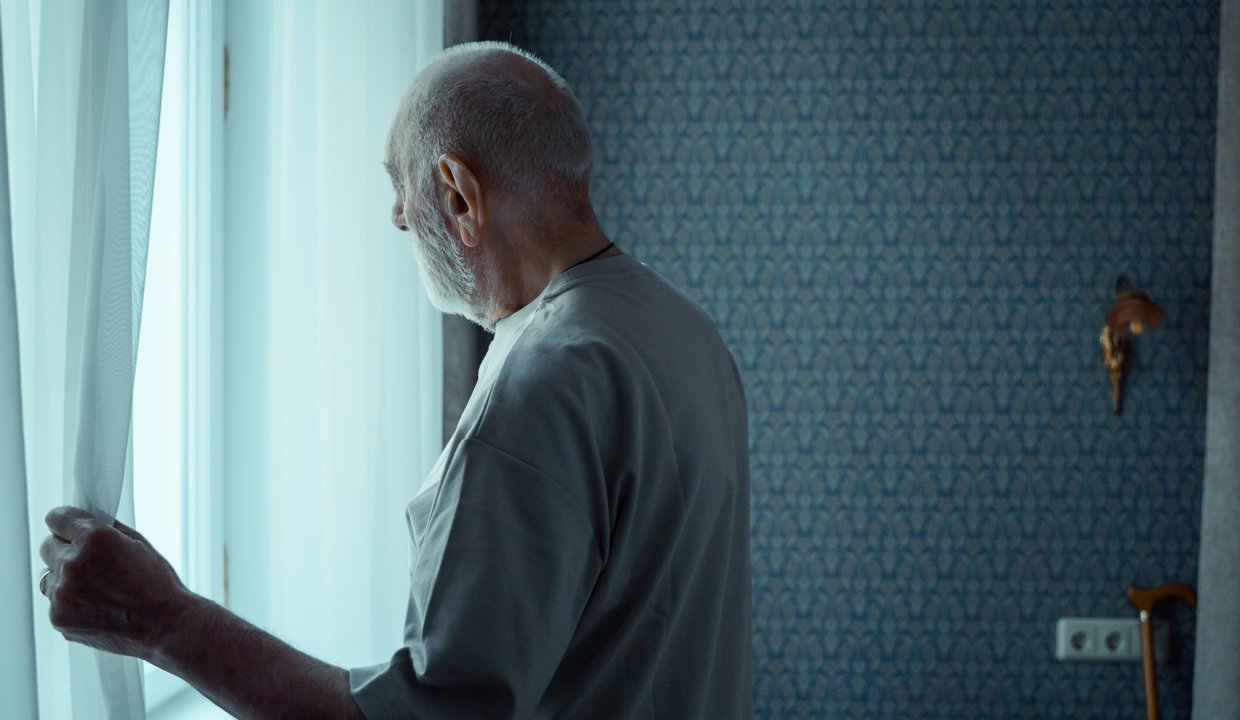 An elderly man looks out of a window after a knock at the door
