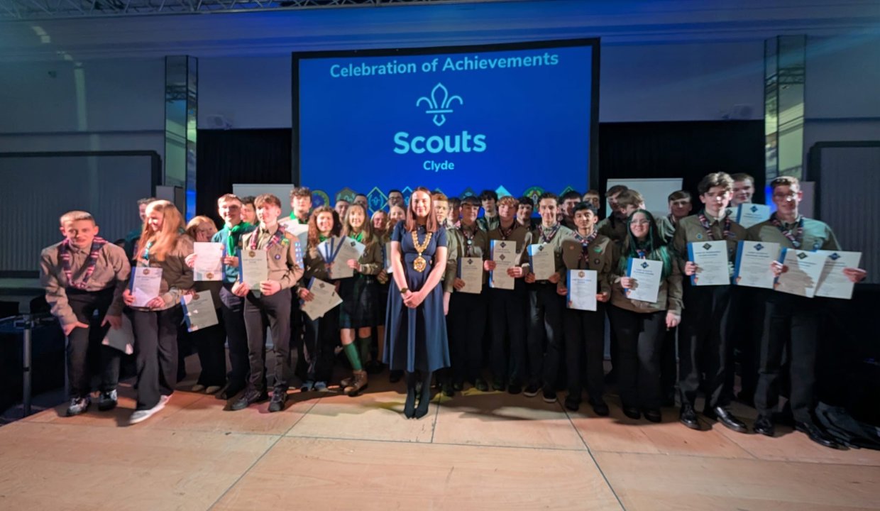 Woman in blue dress wearing chains of office standing in front of at least 50 young people wearing scout uniform holding certificates. Behind them is a back drop with the words ‘Celebration of Achievement – Clyde Scouts’. 