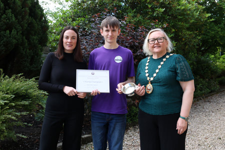 Young man in a purple top standing between two women – the one on the left wearing black and holding a framed certificate, the one on the right wearing a green top, a chain of office and holding a silver quaich. They are outside and there is green foliage behind them. 