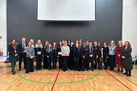 Large group of people standing in a gym hall with a large black wall behind them with a white screen on top. 