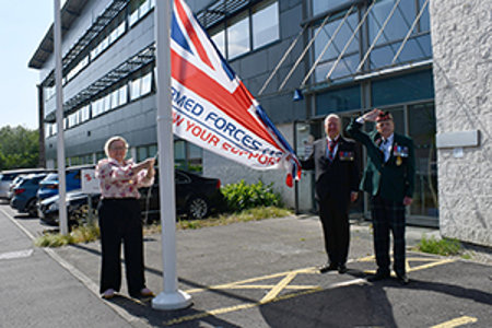 Provost Gillian Renwick, wearing chains of office raises special union flag – the Armed Forces Day Flag. Across from the flag pole stands a man in a suit wearing military medals and a man in military dress, wearing a hat and saluting the raising of the flag. 