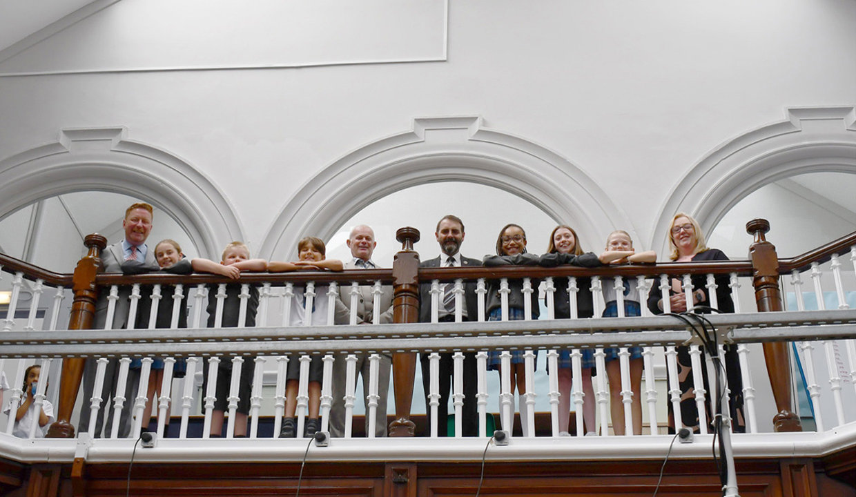 Group of children and adults looking down from a banister in a school. 