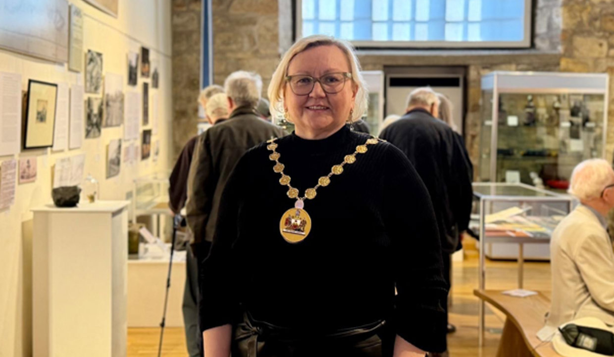 Woman wearing a black top and chains of office standing in large room with exhibits. A brick work wall and large window is behind her. 