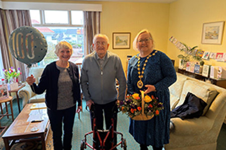 Image of three people standing in bright lemon painted living room, with matching lemon suite. The lady on the far left is holding a blue ‘100’ balloon in her hand.  An older gentleman is standing in the middle with a walking aid and to his right is woman in a blue dress, wearing chains of office and holding a basket of beautiful flowers. 