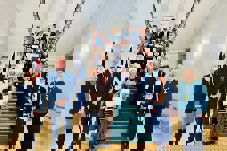 Eighteen men, women and children holding certificates standing on a wide, carpeted staircase.  In the foreground are three people, two of whom are wearing chains of office. Both the Union flag and the Saltire are hanging from a stand on the left of the staircase. 