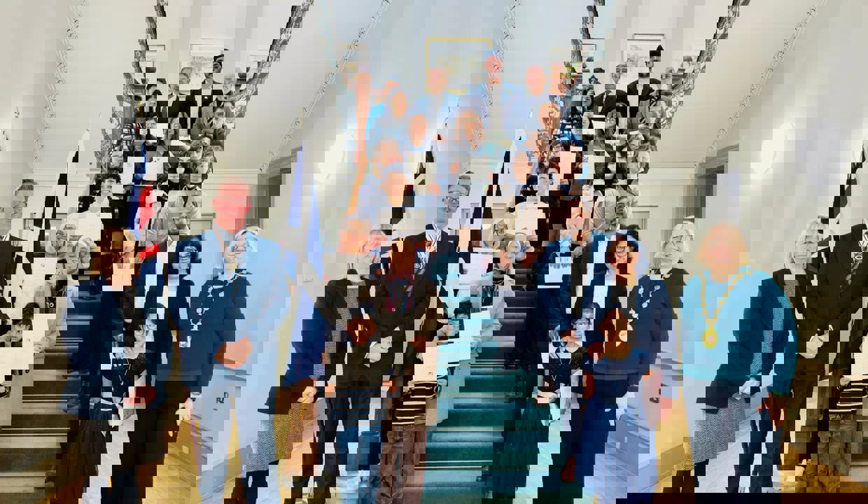 Eighteen men, women and children holding certificates standing on a wide, carpeted staircase.  In the foreground are three people, two of whom are wearing chains of office. Both the Union flag and the Saltire are hanging from a stand on the left of the staircase. 