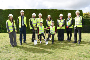 Adults and children wearing hi vis vests and hard hats standing on grass.  A green hedge is behind them. In the foreground is one man and two children digging into the ground with spades. 