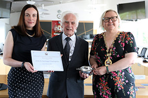 To the left is a woman wearing black, holding a framed certificate next to older gentleman wearing a suit. He is holding one side of a silver quaich and the other side is held by a woman wearing chains of office. 