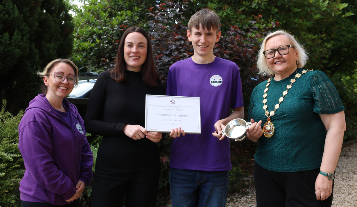 Woman on left wearing purple top beside woman wearing black and holding a framed certificate beside young man in a purple top. To the right of the man is a woman wearing a green top and a chain of office.  She is holding a silver quaich. They are outside and there is green foliage behind them. 