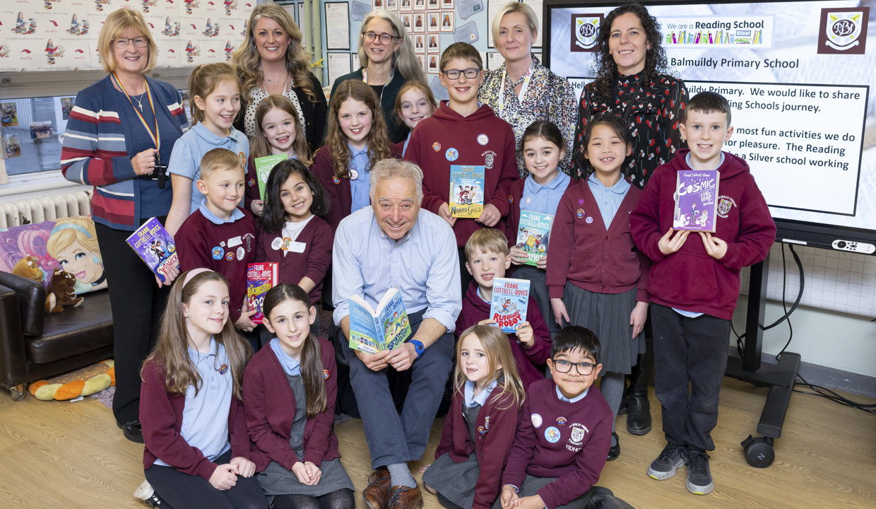 A group of children wearing burgundy school jumpers standing or sitting around a man smiling at the camera and reading a book. Five women are standing at the back of the group, smiling at the camera.