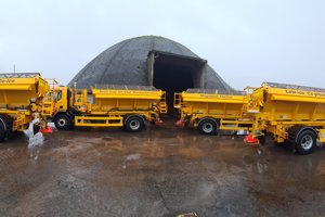 four yellow grit trucks parked in front of dome-shaped salt storage unit