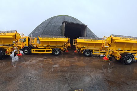 four yellow grit trucks parked in front of dome-shaped salt storage unit