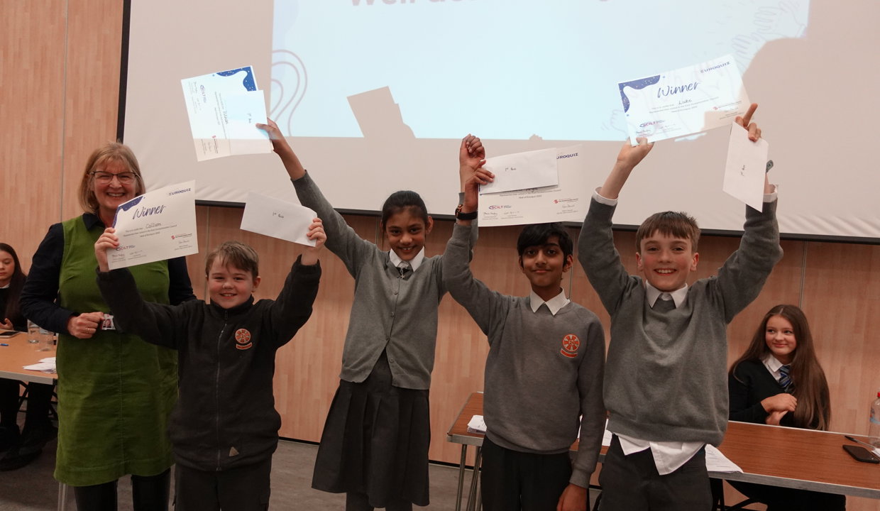 Woman in green dress with four primary school pupils in grey uniform holding ‘winners’ certificates high in the air.