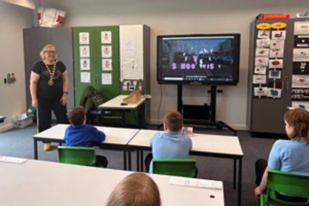 Provost Renwick standing in a classroom at Woodland View School in front of a class of pupils sitting at their desks.  To her left is a tv screen where she is showing her video to the pupils. 