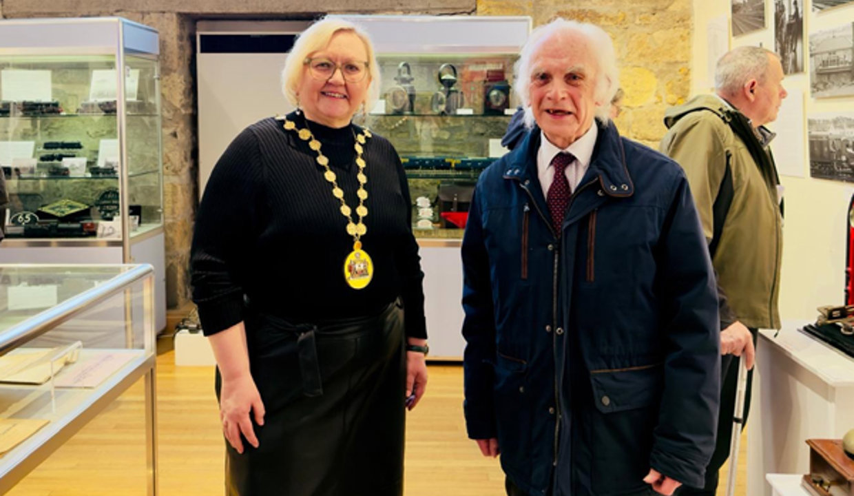 Woman wearing a black top and chains of office standing beside man wearing dark coloured coat and shirt and tie. Behind them is a brick work wall and large window.