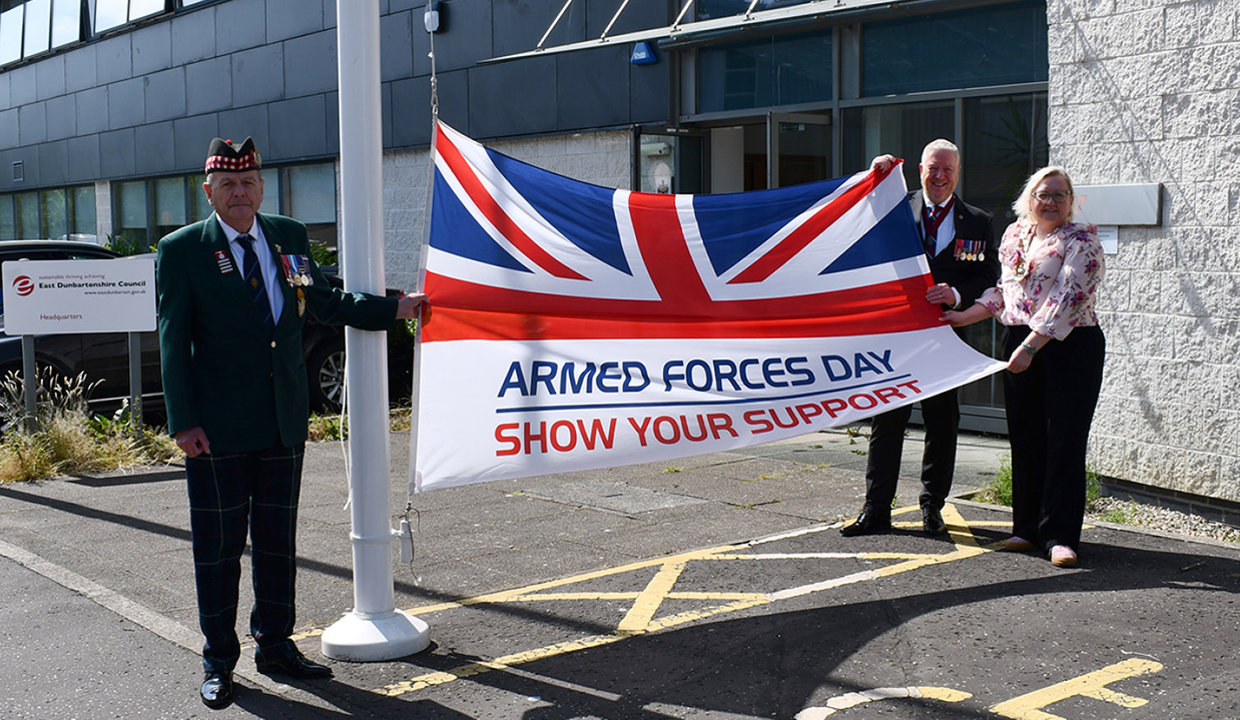 Man in armed forces uniform stands at flagpole holding out the Armed Forces Day Flag which is a special union flag.  The other end of the flag is being held by  a man wearing military medals and Provost Gillian Renwick, wearing the Provost’s chains of office. 